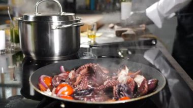 Deniz ürünleri restoranı. Siyah arka planda yükselen buharla tavada ahtapot pişirmek. Siyah eldivenli şef arka planda yemek kesiyor. Restoranın mutfağında yemek pişirmek. Lezzetli büyük ahtapot.