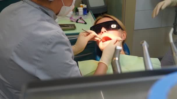 Стоматолог осматривает зубы с помощью медицинских инструментов. Портрет молодой женщины в очках, проверяющей зубы. Пациент с открытым ртом. Лечение зубов. Современная медицинская стоматология. 4 k видео — стоковое видео