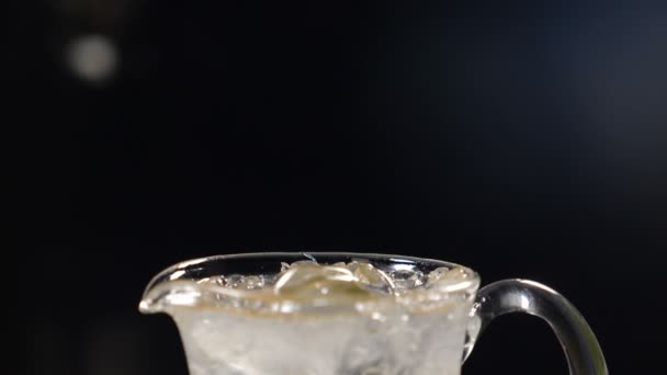 Agua de soda espumosa que se vierte en un frasco de vidrio lleno de lima en rodajas, hojas de menta y hielo, frutas y bayas de verano. Cóctel refrescante sobre fondo negro con cubitos de hielo en la mesa. Movimiento lento — Vídeos de Stock