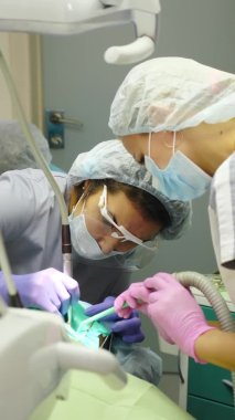 Modern diş kliniği. Dikey video: Tıbbi ekip yeni stomatoloji kliniğinde tedavi sırasında yüz kalkanı, maske ve eldiven takan sterilize aletlerle çalışıyor. Dişçi çocukların dişlerini tedavi ediyor. 4 bin.