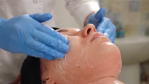 A kozmetikus orvos arctisztító habot használ a női arcon. Kozmetológus készíti a terápiás eljárást. Az orvos kék orvosi kesztyűben kezeli az ügyfelet. Bőrápolás és bőrgyógyászat. arc — Stock videók