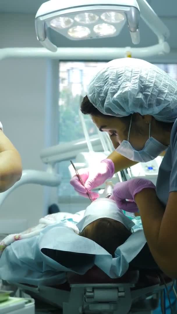 Tratamiento dental en niños bajo anestesia general. El médico dentista trata los dientes de la muchacha. Imágenes verticales. Equipo de cirujanos dentales profesionales que realizan operaciones invasivas en pacientes con — Vídeo de stock