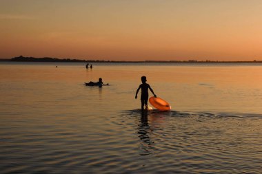 Akşam gölünün arka planında turuncu şişme daire olan çocuk silueti. Turuncu gün batımı. Boşluğu kopyala.