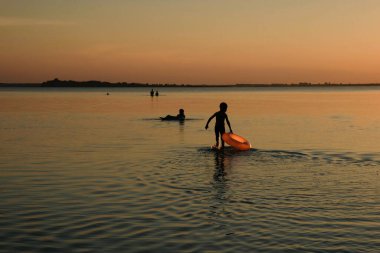 Turuncu şişme daire ve gün batımında bebek silueti. Çocuk sığ suda yürüyor. Boşluğu kopyala.