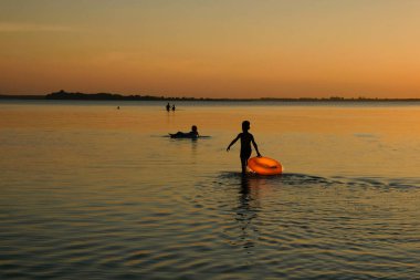 Sahilde günbatımının arka planında insan siluetleri. Bir çocuğun elinde turuncu şişme daire. Boşluğu kopyala.