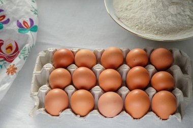 Yumurta kutusunda taze çiftlik tavuğu yumurtası. Kasedeki un. Nakışlı havlu. Paskalya pastası hazırlığı. Sabah ışığı.