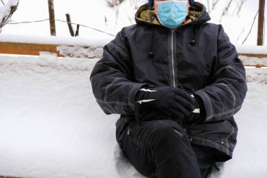 Karla kaplı bir bankta oturan yaşlı bir adam. Koruyucu maske. Sıcak eldivenler. Adam sakin duruyor. Fotoğraf mim 'e benziyor. Boşluğu kopyala.