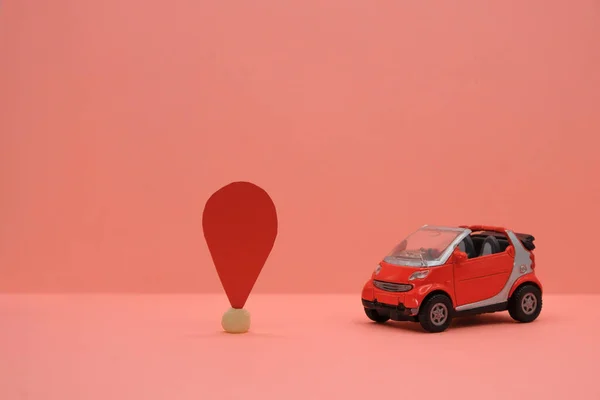 Red small car toy near the location sign. Travel or delivery concept. Car services. Coral background. Copy space.