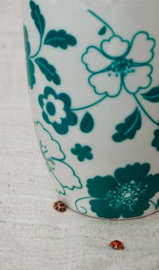 Çiçeklerle süslenmiş bir fincanın yanında gri bir tuval üzerinde uğur böcekleri.