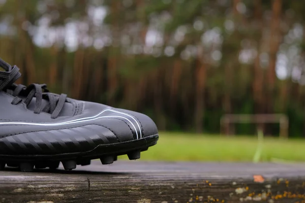 Fragment of black football shoes standing on a wet wooden bench on a football field background. Forest stadium after the rain in Ukraine. Copy space.