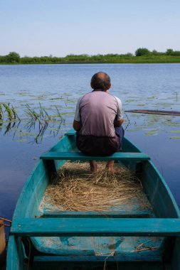 Alone senior sits barefoot in a wooden boat in the river background. Vertical image.