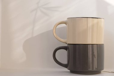 Beyaz arka planda iki boş bej ve gri bardak. Sabah ışığı. Bitkiler duvara yansıyor. Boşluğu kopyala. 