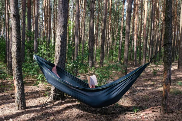 Hammock with a resting man on a pine forest background. A man reads a book while lying in a hammock. Forest camping in Ukraine.