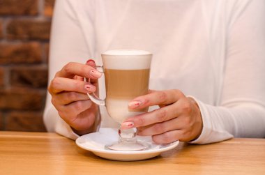 Kafedeki genç kadın sütlü kahve içiyor, elinde şeffaf bir bardakla beyaz bir fincan tabağı tutuyor. Kahvenin soğumasını bekler