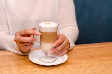 Kafedeki genç kadın sütlü kahve içiyor, elinde şeffaf bir bardakla beyaz bir fincan tabağı tutuyor. Kahvenin soğumasını bekler