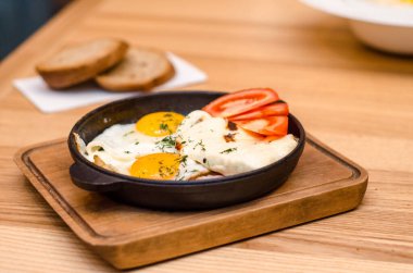 Sıcak kahvaltı siyah tavada servis edilir, pastırmalı ve domatesli sahanda yumurta, ahşap masada ekmek.