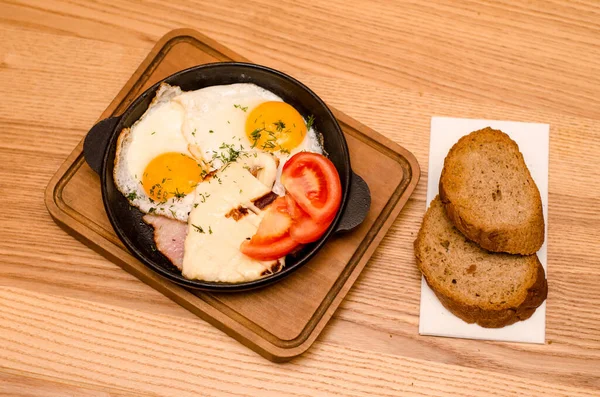 Sıcak kahvaltı siyah tavada servis edilir, pastırmalı ve domatesli sahanda yumurta, ahşap masada ekmek.