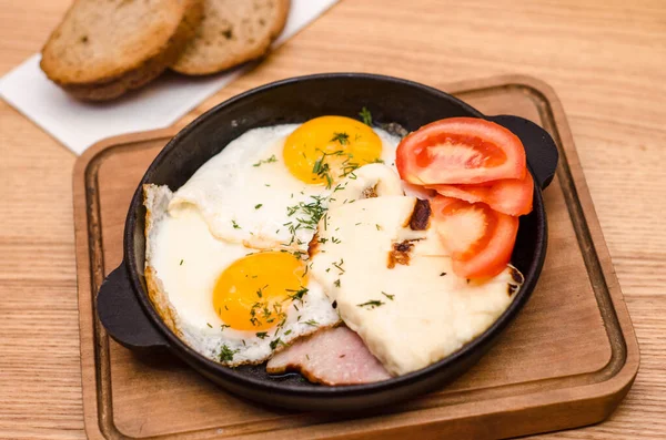 Sıcak kahvaltı siyah tavada servis edilir, pastırmalı ve domatesli sahanda yumurta, ahşap masada ekmek.