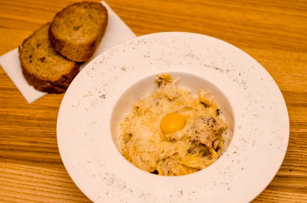 Lokantadaki beyaz tabakta yumurta sarısı ile iştah açıcı carbonara makarnası, ahşap masada iki parça ekmek.