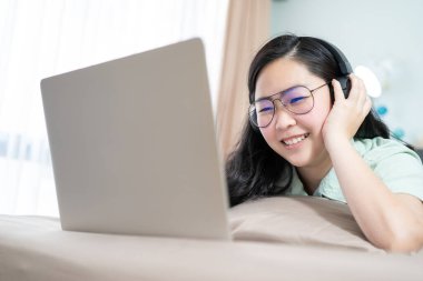 Güzel bir Asyalı kadın dizüstü bilgisayarlı bir kulaklık kullanıyor ve yatak odasındaki yatağa uzanmış kahverengi renkli bir tema kullanıyor..