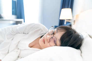 Asyalı genç bir kadın beyaz temiz bir yatakta beyaz tişörtle uzun kollu pijamalarıyla ve gözlerinde gözlük olmasına rağmen uyuyor..