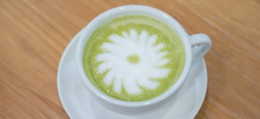 Sıcak bir fincan kibrit sütlü kahve, beyaz bir fincan üzerinde duruyor. Beyaz köpüklü, ortasında basit bir çiçek deseni oluşturan bir tabağın üzerinde..