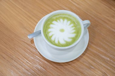 Sıcak bir fincan kibrit sütlü kahve, beyaz bir fincan üzerinde duruyor. Beyaz köpüklü, ortasında basit bir çiçek deseni oluşturan bir tabağın üzerinde..