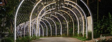 A curved tunnel lined with warm lights glows softly in the dark, and the illuminated arches guide the walkway through a quiet night scene.