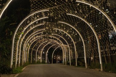 A lit tunnel forms a welcoming route for visitors during nighttime, with rows of warm lights outlining the curved path through the garden.