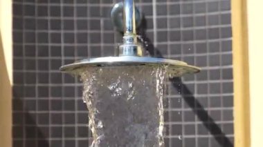 Su yuvarlak metal bir duş başlığından fayanslı bir duvara doğru akar, güneş ışığı su dokusunu ve açık havayı aydınlatır..