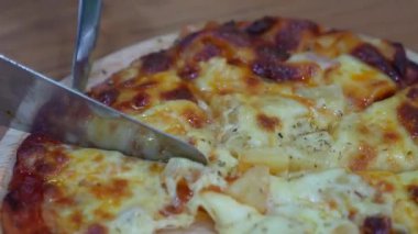 Bir insan, üzerinde erimiş peynir ve ananas parçaları olan yuvarlak bir tahta üzerinde ince bir dilim pizzayı kesmek için bıçak kullanır..