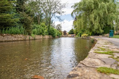 Bourton-on-the-Water, Birleşik Krallık, 12 Temmuz 2023, Ziyaretçiler temiz sığ suların yanında nehir kenarında yürüyorlar. Sahne, parlak yaz gökyüzü altında sakin hareketler gösteriyor..