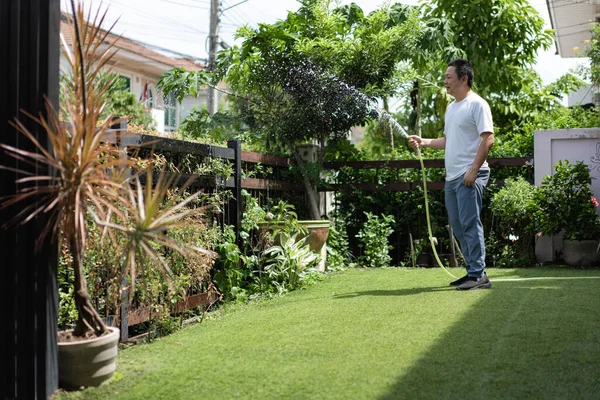 Bir adam güneşli arka bahçesinde rahat bir gülümsemeyle bitkileri sular. Sabah ışığı ve yeşil yapraklar tazeliği ve huzuru ifade eder..
