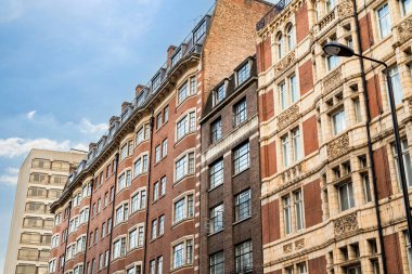 Klasik İngiliz mimarisi, kırmızı tuğlalı binalar ve bulutlu gökyüzünün altındaki tarihi cepheleriyle geleneksel bir Londra caddesini çiziyor..