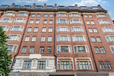 Klasik İngiliz mimarisi, kırmızı tuğlalı binalar ve bulutlu gökyüzünün altındaki tarihi cepheleriyle geleneksel bir Londra caddesini çiziyor..