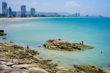 Hua Hin, Tayland 'daki sakin sahil manzarası, ufukta gökdelen manzarası ve mavi gökyüzü. Plaj kayaları ve tropikal bitkiler.