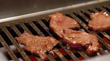 Biftek, görünür ısı ve kahverengi yüzeyi olan metal bir ızgarada pişiriliyor. Bu da profesyonel bir restoran ortamını ve aktif gıda hazırlama sürecini yansıtıyor..