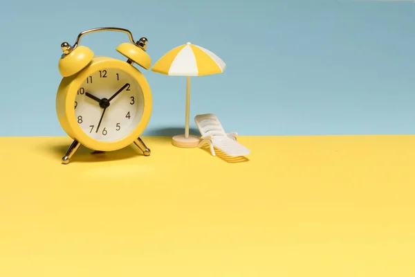 Time to travel. Sun lounger, yellow umbrella and alarm clock on a yellow, blue background.