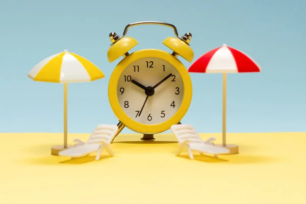 Time to travel. Sun lounger, yellow umbrella and alarm clock on a yellow, blue background.