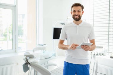 Ofiste tablet kullanan ceketli çekici genç dişçi kameraya bakıyor.