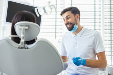 Gülümseyen erkek dişçi klinikte kadın hastayla konuşuyor.