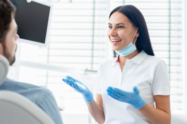 Modern klinikte hastaya tavsiye veren ve el kol hareketi yapan neşeli kadın dişçi.