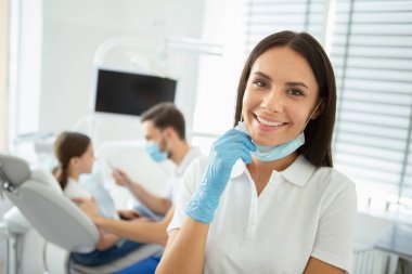 Gülümseyen kadın doktor kameraya bakarken meslektaşı arka planda dişçi koltuğunda çalışan kızla çalışıyor.