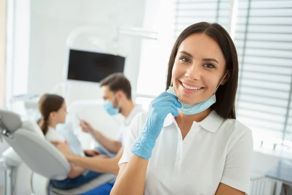微笑的女医生看着摄像机 而她的同事则以坐在牙科椅子上的女孩为背景 — 图库照片