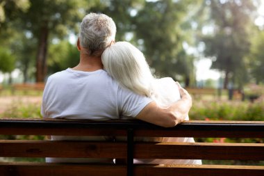 Olgun gri saçlı kadın parkta bir adamın omzuna başını koyuyor.