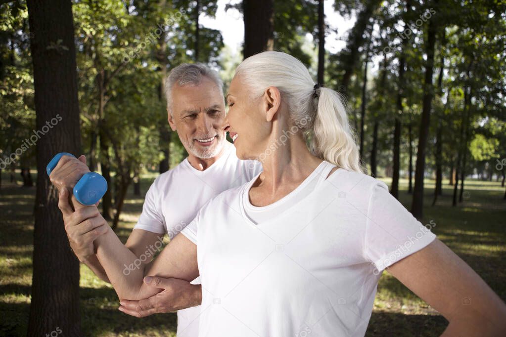 Cheerful man touching the elbow of a mature woman and helping her with hand weight exercising