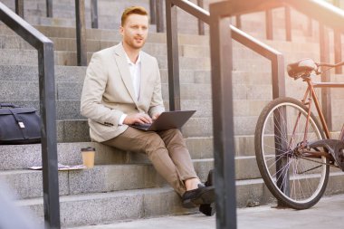 Klasik takım elbiseli karizmatik bir adam laptopla çalışıyor ve merdivenlerde otururken düşünceli davranıyor.