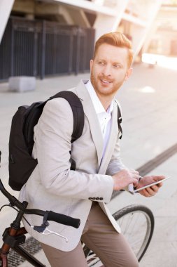 Şık takım elbiseli memnun bir adam bisikletine yaslanmış ve elinde bir tablet tutuyordu.