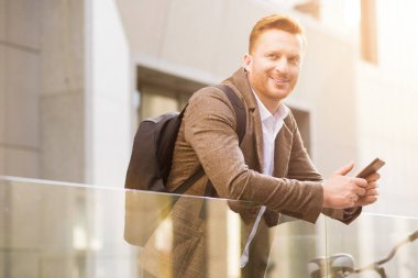 Pozitif stil sahibi bir adam yana bakıyor ve cam panele yaslanırken bir cep telefonu tutuyor.