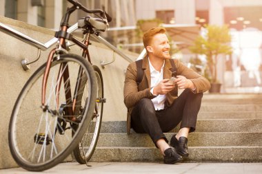 Neşeli, modaya uygun bir adam yana bakıyor ve elinde akıllı bir telefonla dışarıda merdivenlerde oturuyor.